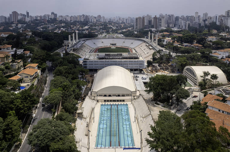 Imagem aérea de um complexo esportivo que inclui uma piscina olímpica, um estádio e áreas verdes ao redor. O estádio está ao fundo, com arquibancadas visíveis, e a piscina está em primeiro plano, com água azul. A cidade se estende ao fundo, com prédios e árvores.