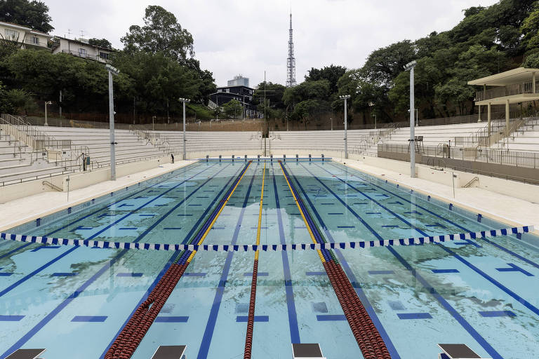 Imagem de uma piscina olímpica com várias raias visíveis. O fundo apresenta arquibancadas vazias e vegetação ao redor. O céu está nublado e há uma torre de transmissão ao fundo.