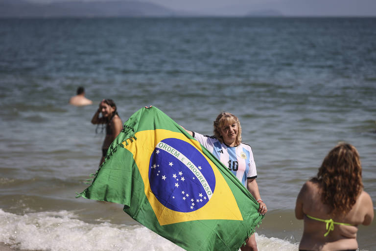 mulher branca loira com camiseta da argentina e canga com bandeira do Brasil em frente a mar é fotografada por amiga de costas de biquini
