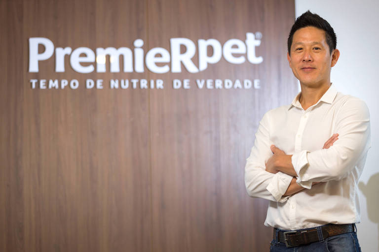 Um homem está posicionado em frente a uma parede com o logotipo da PremieRpet. Ele está vestido com uma camisa branca e tem os braços cruzados. O fundo é de madeira clara e o logotipo é em letras brancas, com a frase 'TEMPO DE NUTRIR DE VERDADE' logo abaixo.