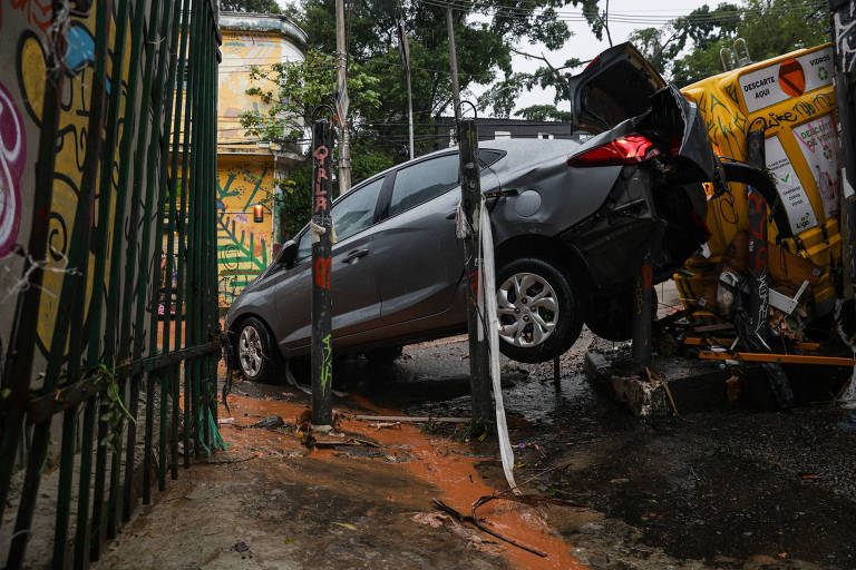 A imagem mostra um carro cinza parcialmente suspenso， com a parte traseira elevada， em uma área urbana. O veículo está próximo a uma calçada e cercas， com um fundo de grafite colorido. Há um veículo amarelo ao lado， e o chão está molhado， possivelmente devido à chuva.