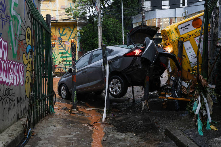 A imagem mostra um carro cinza parcialmente apoiado em um veículo amarelo tombado em uma rua estreita. O carro cinza está em uma posição inclinada, com a parte traseira elevada. Ao fundo, há uma parede com grafites coloridos e vegetação ao redor.