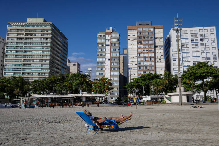 A imagem mostra uma praia com uma pessoa deitada em uma cadeira de sol azul. Ao fundo, há vários edifícios altos e modernos, com árvores na frente. O céu está limpo e azul, indicando um dia ensolarado.