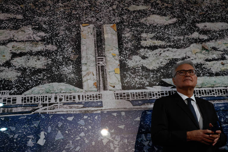 Um homem de terno escuro está em pé em frente a um mural que retrata uma cena arquitetônica com torres e um céu estrelado. O mural é predominantemente em tons de azul e branco， com detalhes que sugerem uma construção. O homem parece pensativo， com as mãos unidas na frente do corpo.
