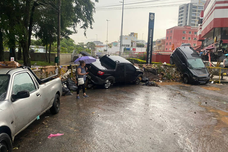 A imagem mostra uma cena de acidente de trânsito em um dia chuvoso. Vários carros estão envolvidos， incluindo um carro preto que está parcialmente virado e um caminhão na posição vertical. Uma pessoa está de pé， segurando um guarda-chuva， enquanto a água da chuva se acumula no chão. Ao fundo， há prédios e uma sinalização visível.