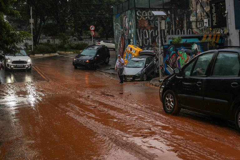 A imagem mostra uma rua alagada com lama， onde vários carros estão estacionados e um veículo está parcialmente submerso. Uma pessoa caminha pela rua， que apresenta marcas de pneus na lama. Ao fundo， há um muro com grafites e árvores ao lado da via.