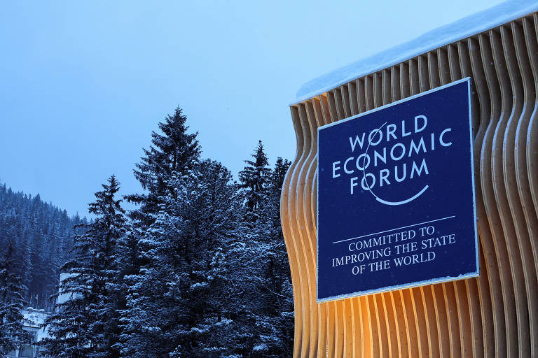 A imagem mostra um painel do Fórum Econômico Mundial, localizado em um ambiente coberto de neve. O painel é de madeira com um design ondulado e contém o texto 'WORLD ECONOMIC FORUM' na parte superior e 'COMMITTED TO IMPROVING THE STATE OF THE WORLD' na parte inferior. Ao fundo, há árvores cobertas de neve, sugerindo um cenário montanhoso e invernal.