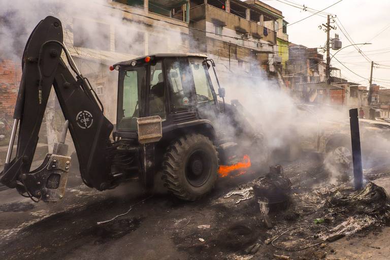 Uma retroescavadeira está operando em uma área urbana， cercada por fumaça. O veículo está em movimento， com a lâmina levantada， enquanto o fundo mostra edifícios e fios elétricos. O ambiente parece caótico， com detritos e fumaça no ar.