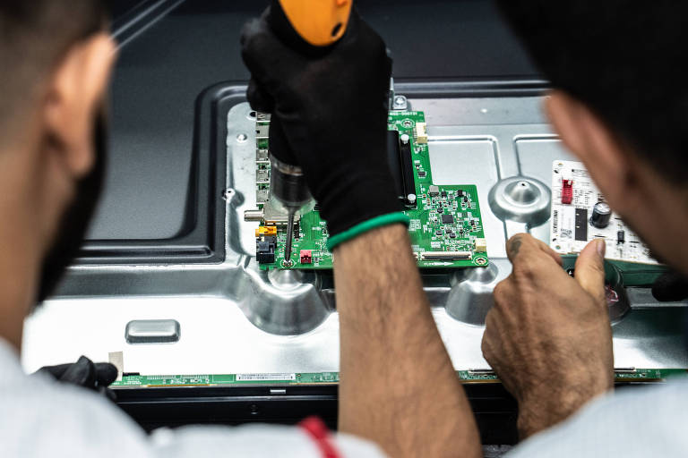 A imagem mostra duas pessoas trabalhando em uma placa eletrônica. Uma delas está segurando uma furadeira ou parafusadeira， enquanto a outra está ajustando componentes na placa. Ambas estão usando luvas pretas e estão concentradas na tarefa. A placa possui diversos componentes eletrônicos visíveis， como resistores e conectores.