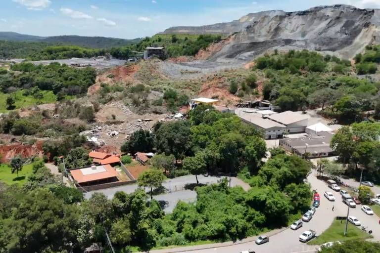 A imagem mostra uma vista aérea de uma área de mineração， com uma grande pedreira ao fundo e vegetação ao redor. A pilha de rejeitos está em parte desabada， atingindo casas. No primeiro plano， há construções e estradas sinuosas， com veículos estacionados. A paisagem é montanhosa， com árvores e colinas visíveis.