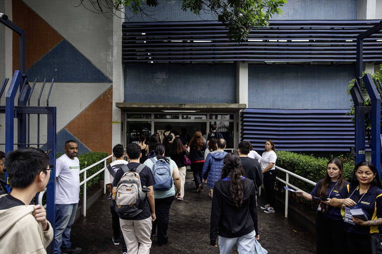 Imagem mostra vários alunos de costas entrando por um portão de universidade. Ao fundo， aparece uma parede azul. Do lado， duas alunas estão paradas， olhando de frente