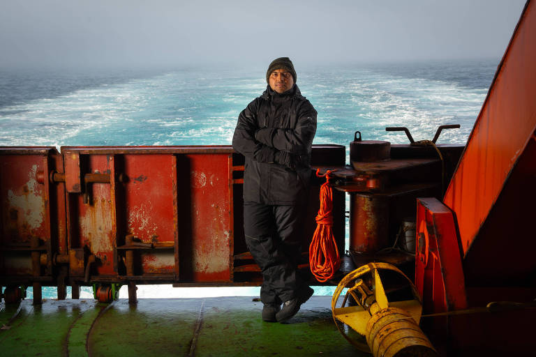 Um homem está em pé em um navio， com os braços cruzados， vestindo roupas escuras e um gorro. Ao fundo， é possível ver o mar e as ondas， enquanto a parte do navio é composta por estruturas metálicas vermelhas e amarelas. O ambiente parece nublado e úmido.