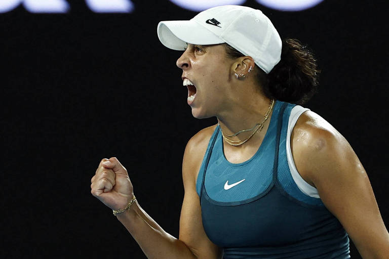 Uma jogadora de tênis está expressando emoção intensa， com um grito de celebração. Ela usa uma camiseta azul e um boné branco. Sua mão está cerrada em um punho， demonstrando determinação e entusiasmo. O fundo é escuro， destacando a figura da atleta.
