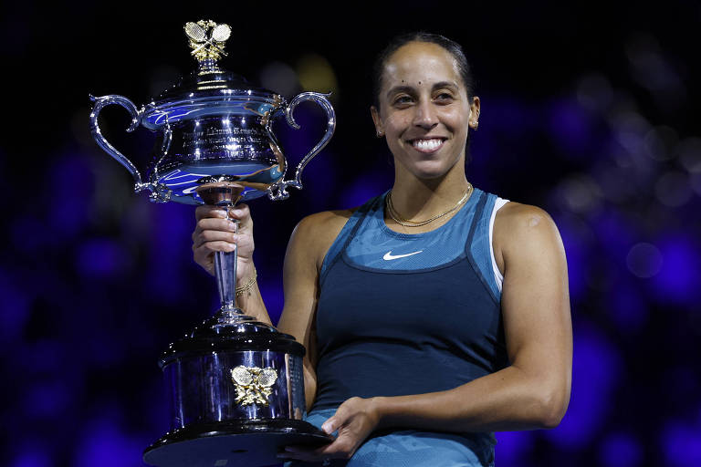 Uma jogadora de tênis está sorrindo enquanto segura um troféu em uma mão. Ela usa uma blusa sem mangas azul e tem cabelo preso. O fundo é desfocado， com luzes roxas e azuis， sugerindo um ambiente de celebração.
