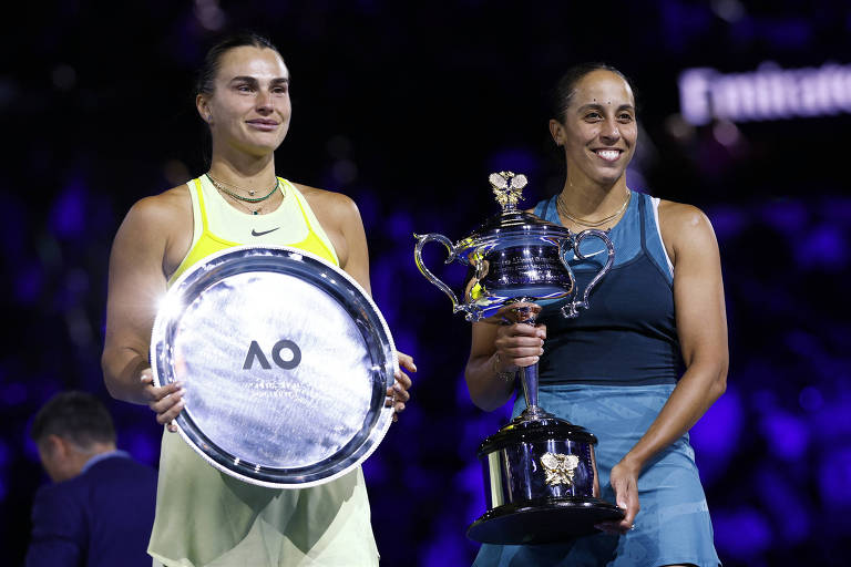 Duas jogadoras de tênis estão em um pódio， uma segurando um troféu e a outra uma placa. A jogadora à esquerda， com um vestido amarelo， exibe uma expressão contida. A jogadora à direita， vestindo um vestido azul， segura um troféu com uma figura em cima e exibe um sorriso. O fundo é iluminado com luzes coloridas.
