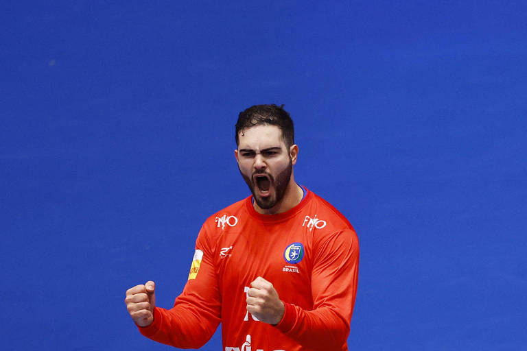 Um jogador de futsal， vestindo um uniforme vermelho， está comemorando com os punhos cerrados e gritando de alegria. O fundo da imagem é azul， destacando a intensidade da sua expressão.
