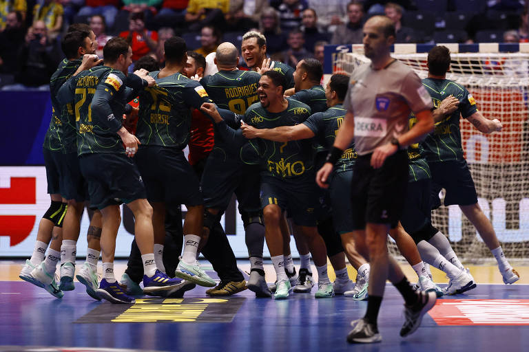 Um grupo de jogadores de handebol celebra em campo， cercando um jogador que parece ter feito um gol. Eles estão vestidos com uniformes verdes e amarelos， e a atmosfera é de alegria e comemoração. Um árbitro está visível no primeiro plano， observando a cena.
