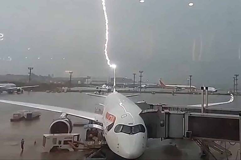 A imagem mostra um avião estacionado em um aeroporto sob um céu escuro e tempestuoso. Um raio é visível descendo em direção ao avião， que está próximo ao terminal. O ambiente ao redor é de chuva intensa， e outros aviões podem ser vistos ao fundo