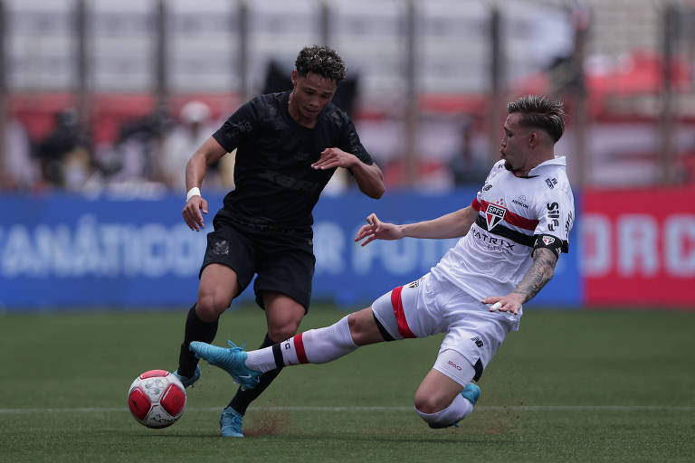 Dois jogadores de futebol estão em campo. Um jogador， vestido com um uniforme preto， está driblando enquanto o outro， vestido com um uniforme branco e vermelho， tenta desarmá-lo com um carrinho. A bola de futebol está próxima ao pé do jogador em uniforme preto. O fundo mostra uma arquibancada com torcedores e banners.