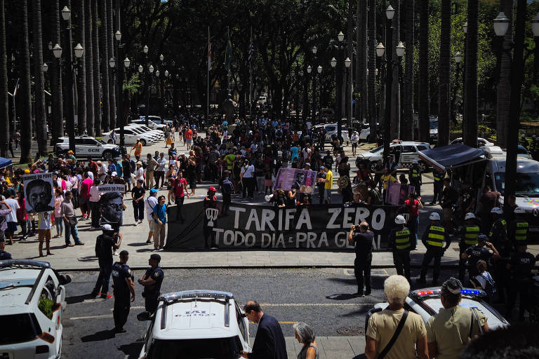 A imagem mostra um grande grupo de pessoas reunidas em uma praça， participando de um protesto. No centro， há uma faixa preta com as palavras 039;TARIFA ZERO TODO DIA É PRA GERAL039;. Algumas pessoas seguram cartazes， e há um forte aparato policial visível， com agentes em uniformes e viaturas. Ao fundo， palmeiras e prédios podem ser vistos， indicando um ambiente urbano.