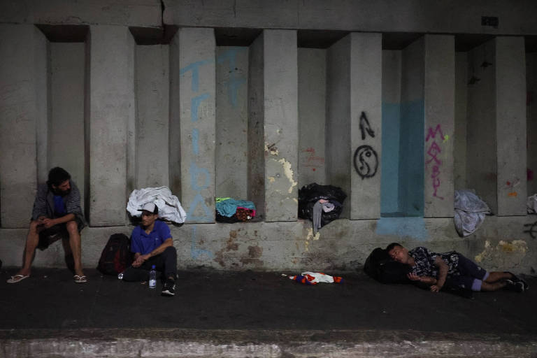 A imagem mostra várias pessoas em situação de rua， sentadas e deitadas sob uma estrutura de concreto. O ambiente é escuro， com paredes de concreto e algumas marcas de grafite. Algumas pessoas estão com mochilas e cobertores， enquanto outras parecem estar descansando no chão.