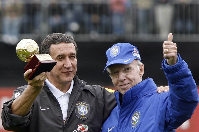 Na imagem， dois homens estão em um evento relacionado ao futebol. O homem à esquerda segura um troféu em forma de bola de ouro， enquanto o homem à direita faz um gesto de positivo com o polegar. Ambos estão vestidos com roupas que representam a seleção brasileira de futebol， com o logotipo da CBF visível. O fundo mostra uma multidão， sugerindo um evento esportivo.