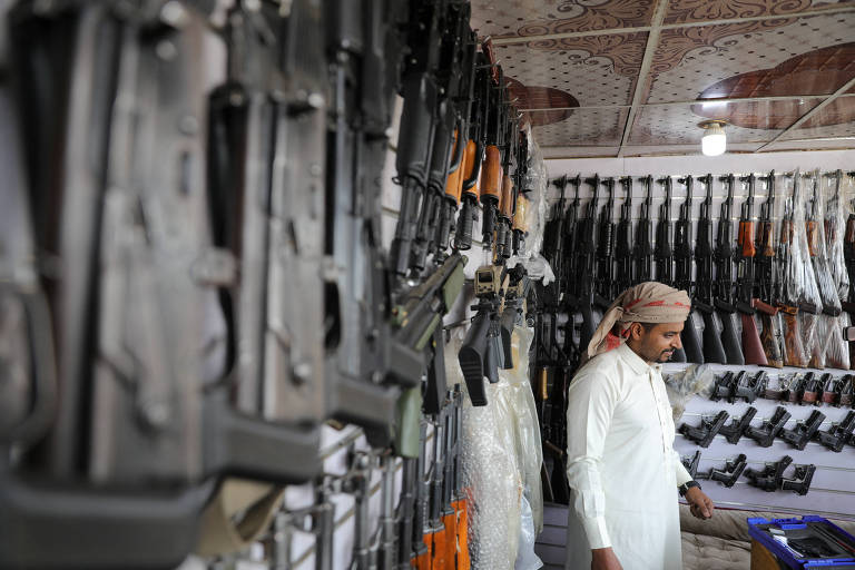 Homem de roupa branca e turbante vermelho e branco entre diversas armas， penduradas na parede