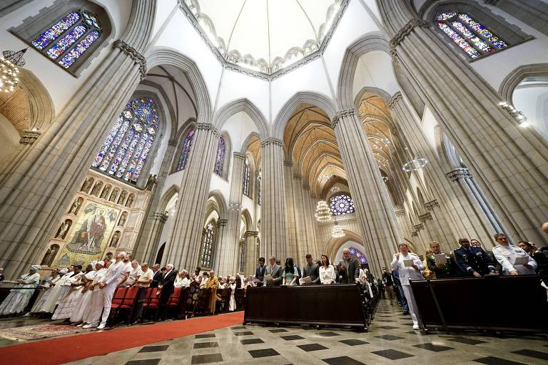 A imagem mostra o interior de uma catedral com grandes colunas e um teto alto. Há um grupo de pessoas em pé， algumas vestindo roupas brancas， em um ambiente iluminado por vitrais coloridos. O chão é de pedras em padrão quadriculado e há um tapete vermelho no centro.
