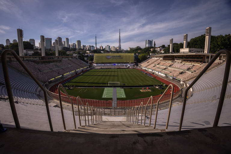 Imagem de um estádio de futebol vazio, com uma vista ampla do campo. As arquibancadas estão em um formato semicircular, e há escadas que descem para o campo. Ao fundo, é possível ver a cidade com prédios altos e torres de comunicação. O céu está claro e ensolarado.