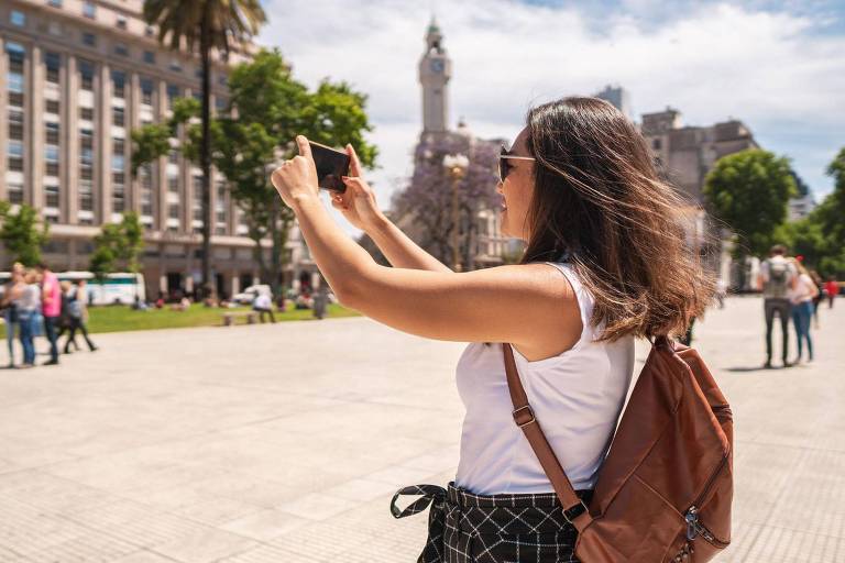 Uma mulher está tirando uma foto com um celular em uma praça. Ela usa uma camiseta branca e uma mochila marrom. Ao fundo， há árvores， edifícios e uma torre visível. O céu está claro e ensolarado.