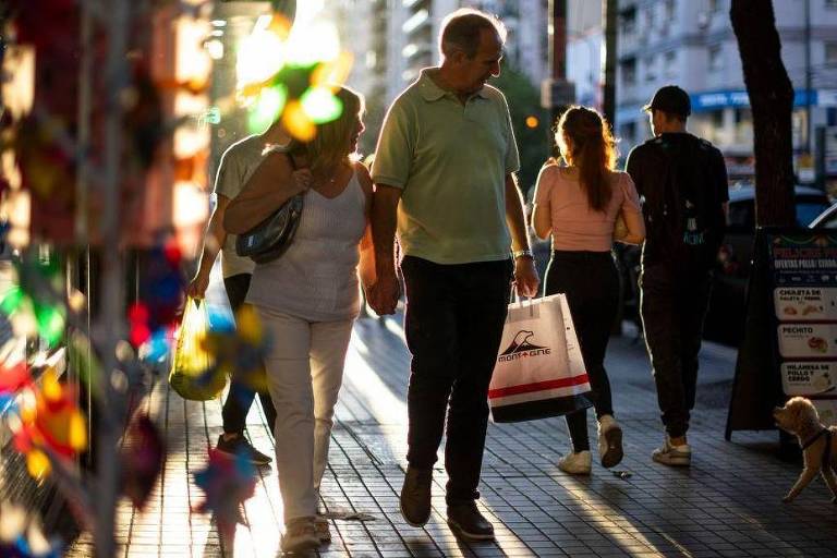 A imagem mostra uma cena de rua durante uma tarde ensolarada. Um homem de cabelos grisalhos está caminhando com uma mulher， ambos segurando sacolas de compras. Ao fundo， outras pessoas também estão caminhando， e há um cachorro pequeno na calçada. A luz do sol cria um efeito de brilho nas superfícies e há elementos coloridos visíveis à esquerda， possivelmente de uma vitrine.