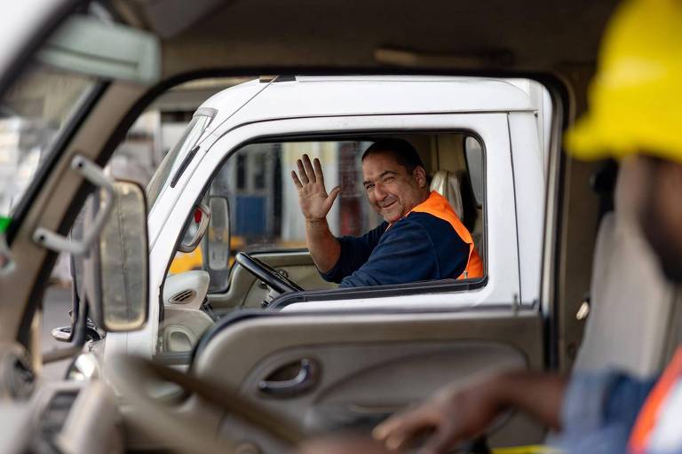 A imagem mostra dois trabalhadores dentro de caminhões. Um deles， à esquerda， está sorrindo e acenando com a mão， enquanto o outro， à direita， usa um capacete amarelo e parece estar focado em algo à sua frente. Ambos estão em um ambiente de trabalho， possivelmente em um canteiro de obras.