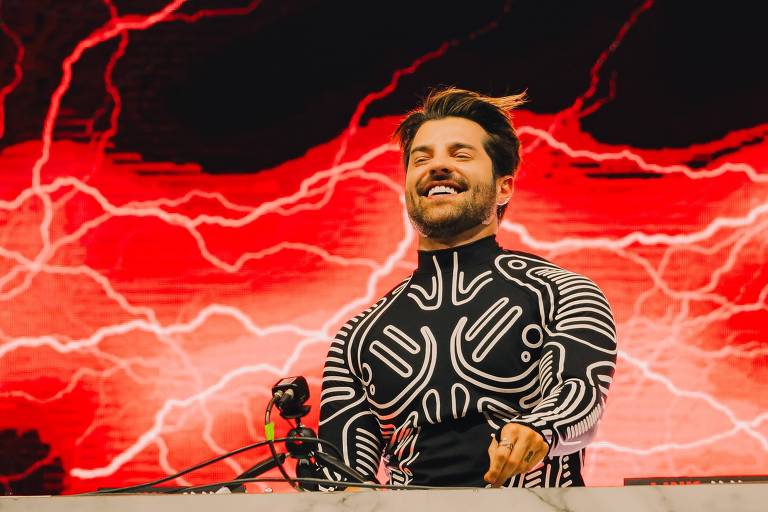 Um DJ está se apresentando em um palco, com um fundo de luzes vermelhas e um céu escuro. O DJ está sorrindo e usando uma roupa com um padrão gráfico. Há uma luz brilhante acima dele, criando um efeito dramático na cena.