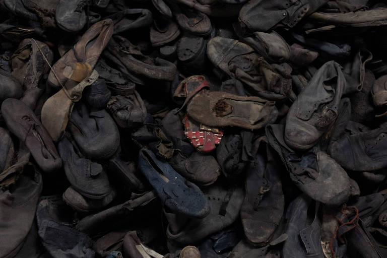 A imagem mostra uma grande quantidade de calçados usados， predominantemente em tons escuros， dispostos de forma desordenada. Entre os sapatos， há um par de sapatos de cor vermelha que se destaca no meio dos outros， que são principalmente pretos e marrons.