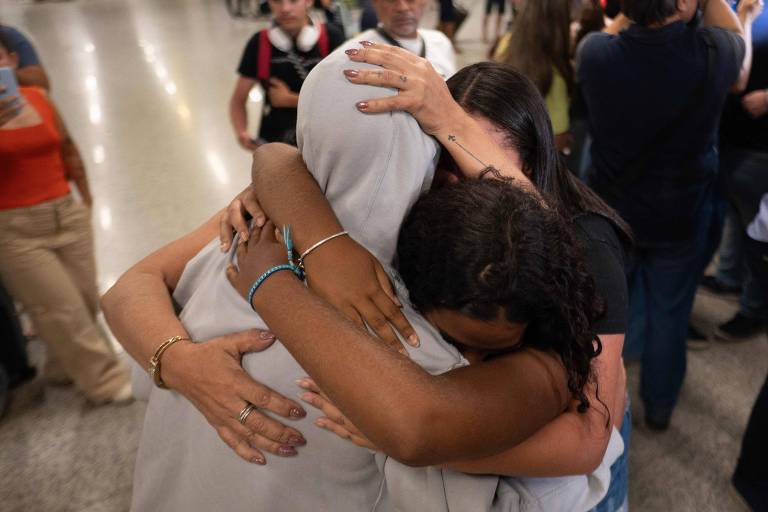 homem vestindo casaco com capuz branco é abraçados por duas mulheres em saguão de aeroporto 