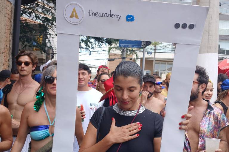 Uma mulher em um traje de banho segura uma moldura grande， que parece ser uma representação de uma rede social. Ela está em um evento ao ar livre， cercada por outras pessoas que também estão vestindo roupas de banho. A moldura tem desenhos e corações， e a mulher está com a mão sobre o coração， demonstrando emoção.