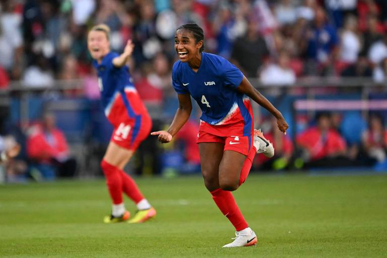 Uma jogadora de futebol， vestindo um uniforme azul e vermelho， está correndo em direção à câmera com um sorriso no rosto， celebrando um gol. Ao fundo， outra jogadora está levantando os braços em comemoração. A torcida está visível nas arquibancadas， com várias pessoas assistindo ao jogo.
