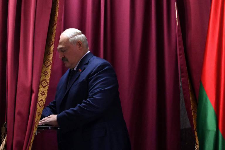 Homem calvo de cabelo branco e bigode escuro， de terno azul escuro， deposita voto em urna. Atrás dele há um pano cor de vinho e uma bandeira vermelha e verde