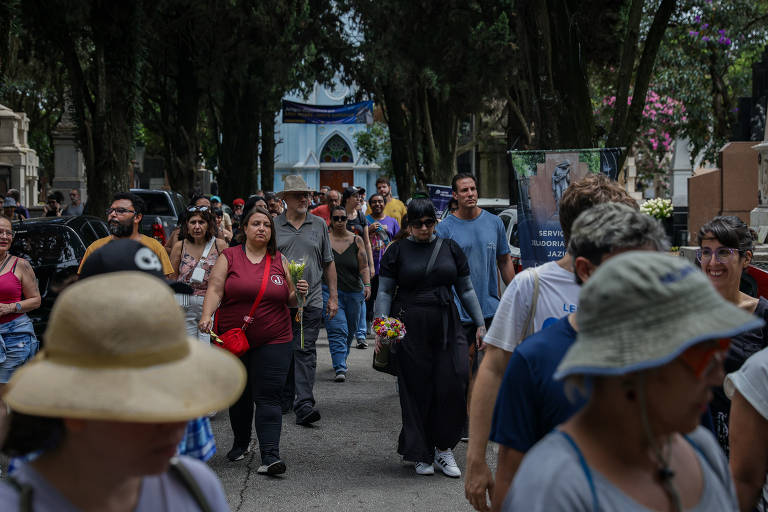 A imagem mostra um grupo de pessoas caminhando em um espaço ao ar livre, cercado por árvores. Algumas pessoas estão usando chapéus, e há uma mulher vestida de preto no centro da imagem. Ao fundo, é possível ver uma construção com uma fachada azul e outros elementos decorativos.