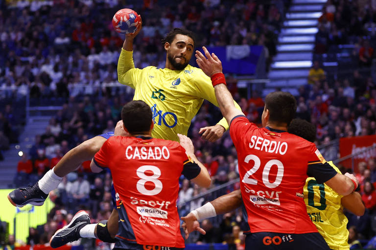 Um jogador da equipe do Brasil， vestido com uma camisa amarela， está saltando e arremessando uma bola de handebol. Dois jogadores da equipe adversária， vestidos com camisas vermelhas， estão tentando bloquear o arremesso. Um deles， identificado como 039;Casado039; com o número 8， está à esquerda， enquanto o outro， identificado como 039;Serdio039; com o número 20， está à direita. O fundo mostra uma plateia com torcedores.