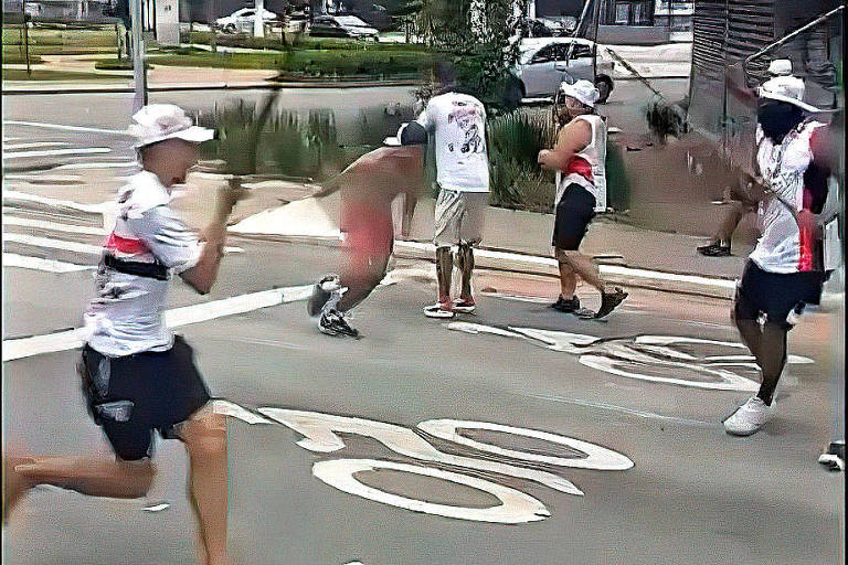 A imagem mostra um grupo de pessoas em uma rua， algumas delas segurando bastões. Um indivíduo parece estar em uma posição defensiva， enquanto outros estão em movimento. O ambiente urbano é visível ao fundo， com carros e vegetação.