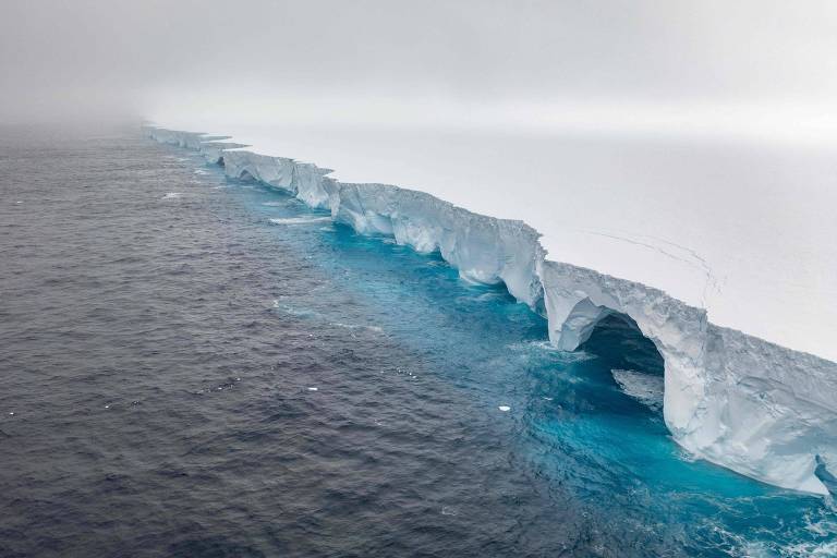 A imagem mostra um iceberg flutuando em águas oceânicas. A parte superior do iceberg é branca e apresenta uma borda irregular， enquanto a parte submersa é visível em um tom azul claro. O fundo é nublado， criando uma atmosfera de neblina ao redor.