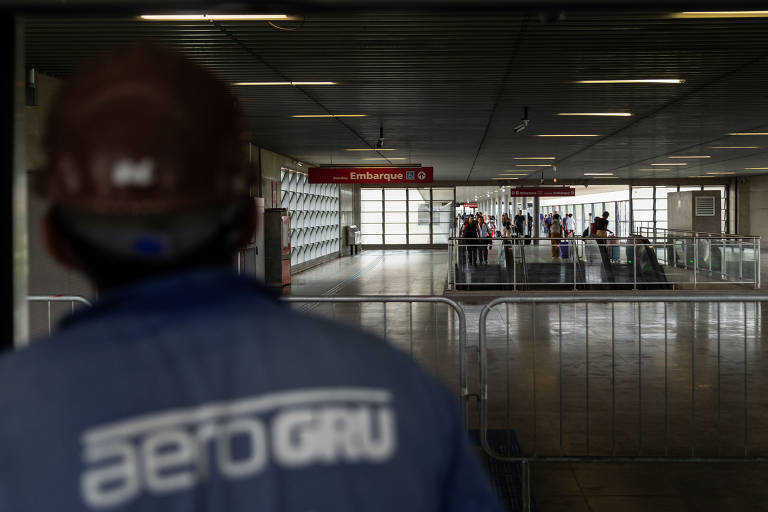 A imagem mostra um funcionário de costas, vestindo uma camisa azul com a inscrição 'aeroGRU'. Ele está em um ambiente de aeroporto, observando uma área de embarque onde várias pessoas estão na fila. Ao fundo, há uma sinalização indicando 'Embarque B'. A iluminação é natural e o ambiente parece movimentado.