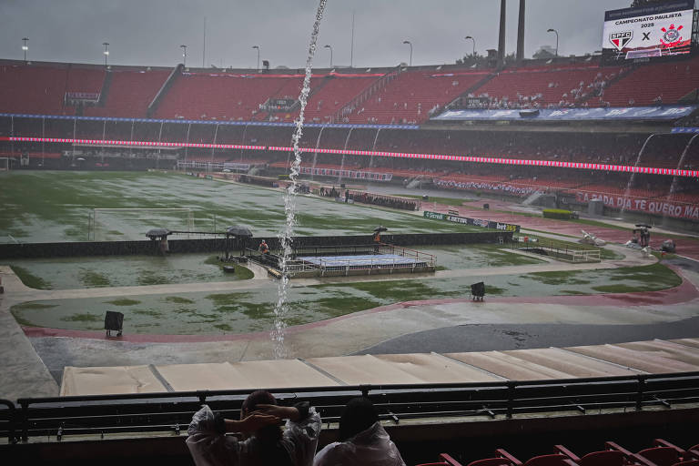 A imagem mostra um estádio de futebol durante uma forte chuva. O campo está alagado， com água acumulada em várias áreas. Algumas pessoas estão sentadas na arquibancada， usando capas de chuva. No fundo， há uma tela eletrônica exibindo informações sobre um jogo entre SPFC e Corinthians. O céu está nublado e a chuva cai intensamente， criando um efeito de água escorrendo em algumas partes do estádio.