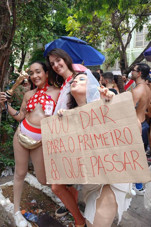 Três mulheres posam para a foto em um evento ao ar livre. Duas delas usam maiô colorido， enquanto a outra está vestida de noiva e segura um cartaz feito de papelão. O cartaz contém texto escrito à mão. Ao fundo， há pessoas e árvores， sugerindo um ambiente festivo.