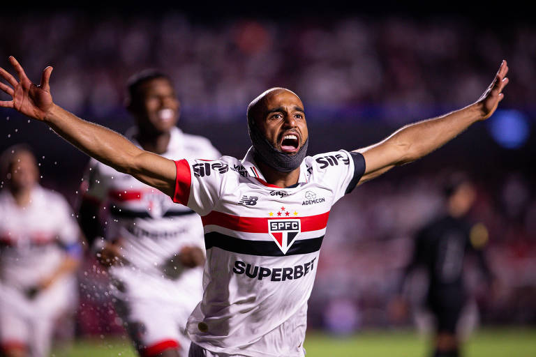 Um jogador de futebol está comemorando um gol， com os braços abertos e gritando de alegria. Ele veste uma camisa branca com detalhes em vermelho e preto， que representa o São Paulo FC. Ao fundo， outros jogadores estão visíveis， e a atmosfera é de celebração. O jogador parece estar em um estádio iluminado， com torcedores ao fundo.