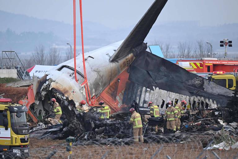 A imagem mostra os destroços de um avião acidentado， com a fuselagem danificada e parte da asa queimada. Equipes de bombeiros e resgate estão trabalhando na cena， utilizando equipamentos para controlar o fogo e investigar o acidente. Ao fundo， é possível ver veículos de emergência e uma área de fumaça