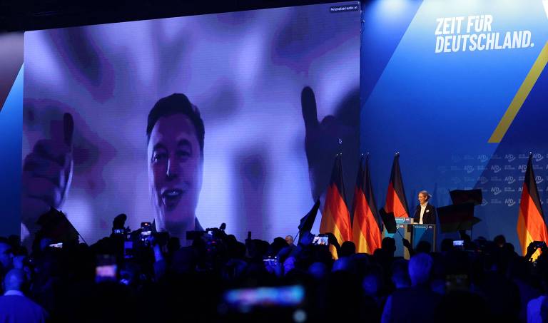 A imagem mostra um grande palco em um evento， onde uma pessoa aparece em uma tela grande， fazendo um gesto com as mãos. O público está em frente ao palco， com várias bandeiras da Alemanha ao fundo. A iluminação é predominantemente azul， criando um ambiente vibrante.