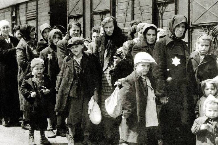 A imagem mostra um grupo de pessoas em fila， possivelmente esperando para embarcar em um trem. A maioria das pessoas está vestida com roupas escuras e algumas usam lenços na cabeça. Entre elas， há crianças， algumas segurando sacolas. Um menino na frente usa um chapéu e uma jaqueta escura. Ao fundo， vê-se vagões de trem.