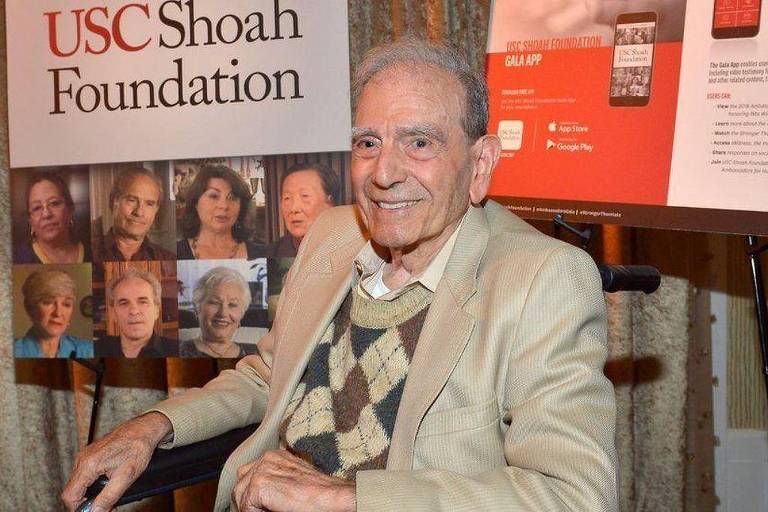 Um homem idoso está sentado em uma cadeira de rodas， sorrindo. Ele usa um suéter claro com um padrão de losangos e uma camisa branca. Ao fundo， há um banner da USC Shoah Foundation com fotos de várias pessoas. Também há um cartaz com informações sobre um aplicativo.